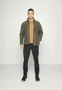 Giacca di pile verde oliva con tasca frontale con zip, indossata sopra una maglia marrone e abbinata a pantaloni neri e sneakers scure.