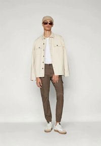 Creme knap-uppen jakke, hvid T-shirt, brune tapered cargo bukser og hvide sneakers. Tilbehør inkluderer en brun beanie og solbriller.