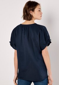 Navy blue short-sleeve blouse with a relaxed fit, pleated back, and gathered accents on sleeves. Smooth texture with subtle sheen.