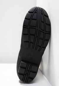 Semelle de chaussure en caoutchouc noir avec des dessins de profondeur et des rainures horizontales et verticales, positionnée contre un mur d'angle blanc.