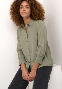 Blusa verde oliva a maniche lunghe con chiusura a bottoni, polsini elasticizzati e texture morbida, abbinata a jeans in denim scuro.