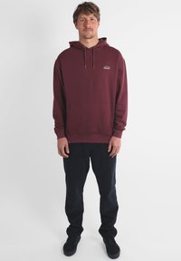 Sweat-shirt maroon avec poche avant et capuche à cordon. Associé à un pantalon noir et des chaussures de sport, sur un fond blanc uni.