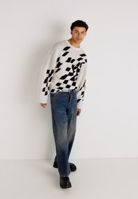 Genser med svart abstrakt mønster på hvit bakgrunn, kombinert med blå jeans. Modellen har på seg svarte klumpete sko og har en avslappet holdning.