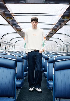 Jonge man met witte trui en donkere spijkerbroek staand in het gangpad van een lege vintagebus met blauwe stoelen en glazen dak.