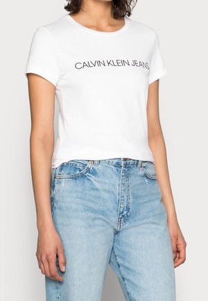 Woman wearing white Calvin Klein Jeans t-shirt and light blue high-waisted jeans, standing against plain background.