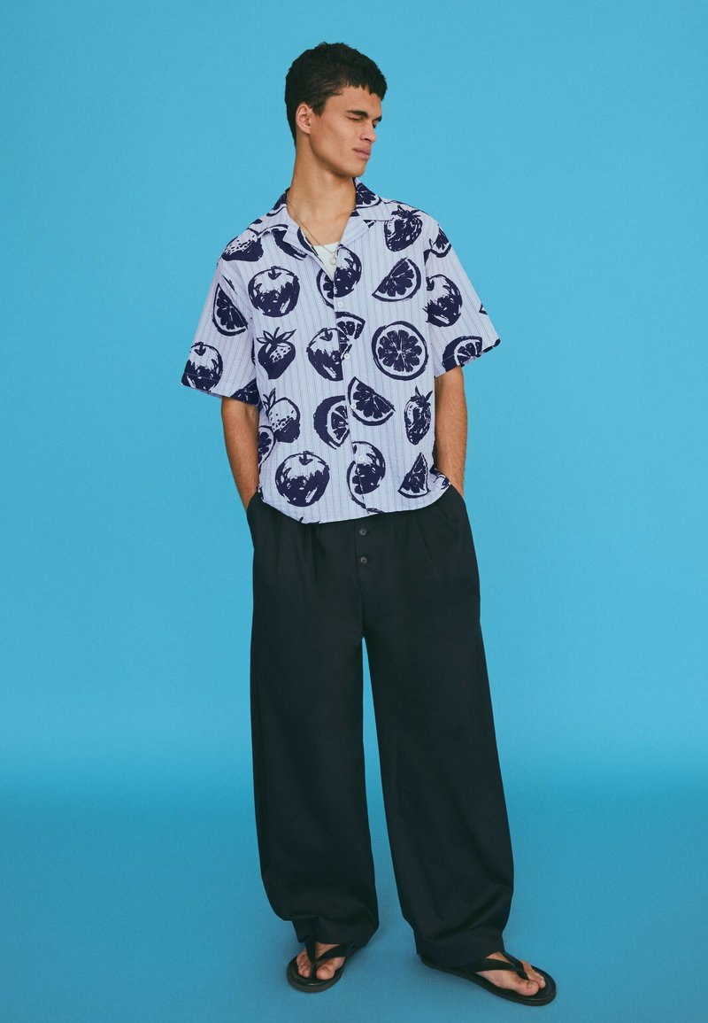 Jeune homme portant une chemise à manches courtes avec imprimé de fruits, pantalon noir ample et sandales, debout les mains dans les poches devant un fond bleu.