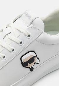 Sneaker bianchi in pelle con superficie testurizzata, dotate di un'emblema a cartone nero e beige e dettagli in argento sul linguetta. I lacci sono bianchi.