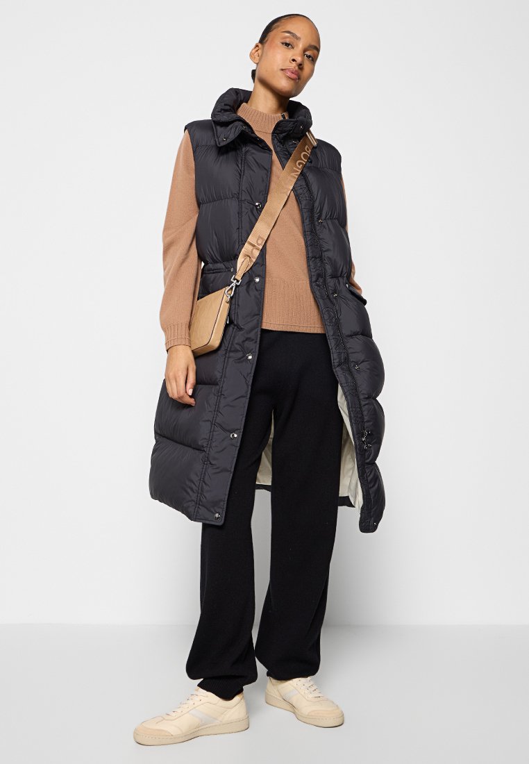 Schwarze gepolsterte Weste mit Kapuze, Druckknöpfen und seitlichen Taschen, getragen über einem braunen Pullover und schwarzen Hosen, kombiniert mit beigen Turnschuhen.