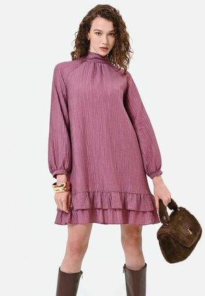 Femme portant une robe mauve à manches longues avec des volants, tenant un sac à main en fourrure marron et portant des bottes hautes marron foncé ainsi que des bracelets en or.