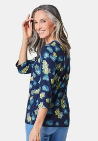 Marineblaue Bluse mit Dreiviertelärmeln, die ein florales Muster in Blau-, Gelb- und Grüntönen aufweist, aus einem weichen, strukturierten Stoff.