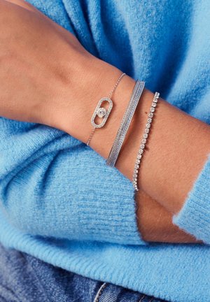 Silverarmband lagda på ett handled: ett med en central rund accent, ett med en strukturerad band och ett med små klara stenar, mot en blå tröja.