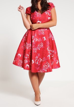 Vrouw die een knie-lange rode bloemenjurk met kapmouwen en witte hoge hakken draagt, staand met gekruiste armen en één hand opgeheven.