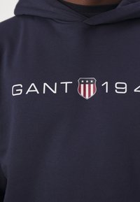 Sudadera azul marino con una textura suave, que presenta letras blancas "GANT" y un logo de escudo con rayas y estrellas rojas y blancas.