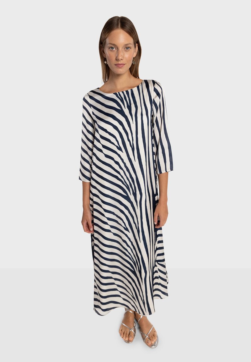 Robe longue ample en ivoire avec des rayures verticales bleu marine. Manches trois-quarts et encolure bateau. Sandales plates visibles.