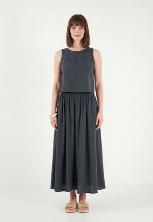 Vrouw met steil bruin haar, gekleed in een mouwloze donkergrijze top en bijpassende lange rok, beige sandalen en armbanden tegen een effen achtergrond.