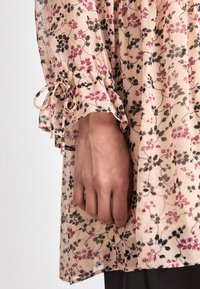 Blomstrete kjole i myk beige stoff med rosa og svarte mønstre. Har innsamlede ermer med sløyfebånd og en avslappet passform.