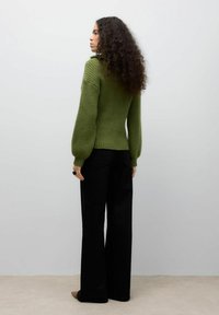 Maglione verde a coste con maniche a palloncino, vita aderente, abbinato a pantaloni neri a gamba larga. Modella vista di spalle, che mostra il retro.