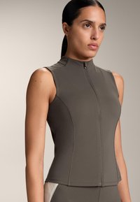 Vrouw draagt een mouwloos grijs jack met een rits en een hoge kraag, staand tegen een effen neutrale achtergrond, kijkt iets naar rechts.