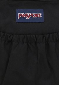 Zwarte stoffen rugzak met een gathered top, voorzien van een blauwe Jansport logo patch met rode letters. Het materiaal lijkt duurzaam en gestructureerd.