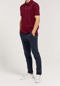 Burgundská polo tričko s krátkým rukávem, bílé logo, v kombinaci s námořnickými chinos kalhotami a bílými teniskami, předvádějící ležérní outfit.
