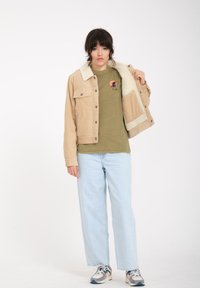 Giacca in corduroy beige con un morbido colletto in pelliccia sintetica crema, abbinata a una maglietta verde con una grafica di palma, jeans dritti azzurri chiari.