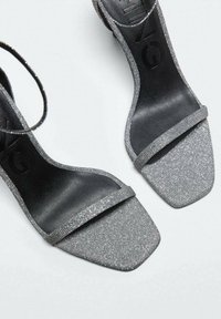 Sandales à paillettes argentées avec un bout carré, semelle noire et fine bride de cheville. Le dessus texturé ajoute une finition scintillante.