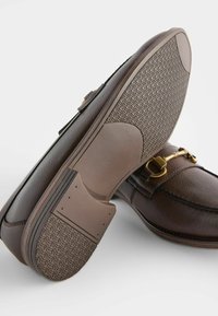 Mocassins en cuir marron avec un extérieur texturé, des accents en matériel doré et une semelle en caoutchouc à motif. Présente une finition lisse et une forme structurée.
