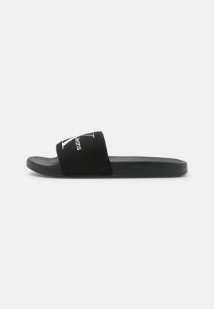 Black slide sandal with cushioned sole and wide fabric strap featuring white "Calvin Klein Jeans" logo on plain background.
