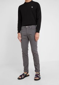 Chemise noire à manches longues avec un petit logo, jean gris ajusté, et sandales rayées multicolores à semelle plate et bout ouvert.