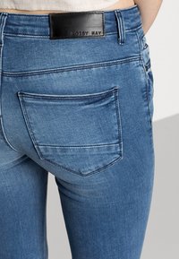 Vaqueros de mezclilla azul con un diseño ajustado y bolsillo trasero visible. Presenta una etiqueta de cuero negro en la cintura marcada con "NOISY MAY".