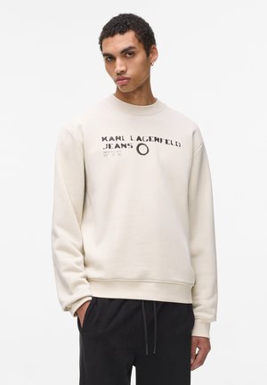 Cream crew-neck sweatshirt with printed black text; long sleeves; relaxed fit; ribbed cuffs; worn with black pants.