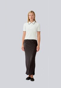 White polo shirt with black scalloped collar, paired with a long black satin skirt. Model stands in flat black shoes. Neutral background.