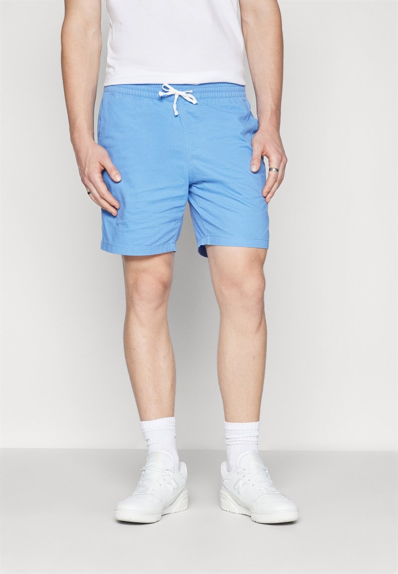 Homme portant un short à cordon bleu clair, des chaussettes blanches, des baskets blanches et un t-shirt blanc, debout devant un fond uni.