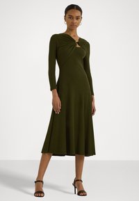Olivgrön klänning med en vriden design vid halsringningen, långa ärmar och en flödande A-linjeformad kjol. Matchad med svarta remsandaler.
