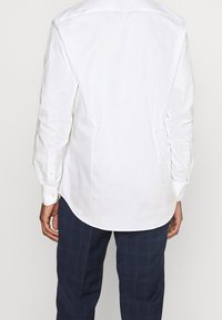 Camicia bianca a maniche lunghe con una texture liscia, caratterizzata da un orlo curvo e polsini con bottoni. Indossata con pantaloni scuri a quadri.
