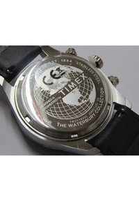 Stainless steel watch back with engraved globe design, "TIMEX," and "THE WATERBURY COLLECTION." Black leather strap visible.