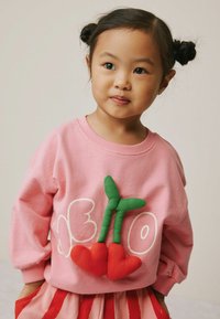 Pink sweatshirt featuring green plant detailing and red heart shapes, with white text. Long sleeves, soft texture. Part of a layered outfit.