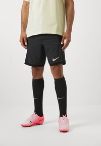 Shorts Nike noirs avec un logo doré, accompagnés de chaussettes noires montantes et de chaussures à crampons roses avec des accents blancs. Design simple et sportif.