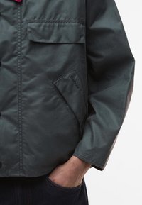 Σκούρο πράσινο μπουφάν με κούμπωμα, πλαινές τσέπες με καπάκι και διακριτική σήμανση σ�την τσέπη. Λείο, αδιάβροχο υλικό.