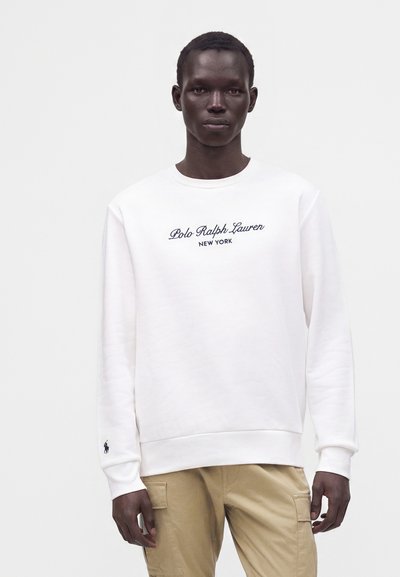 Νέος άνδρας που φοράει λευκό φούτερ Polo Ralph Lauren με σκούρο μπλε κείμενο λογότυπου και μπεζ παντελόνια cargo, στέκεται μπροστά σε απλό ανοιχτό φόντο.