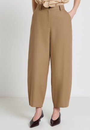 Donna che indossa pantaloni cammello a vita alta e gamba larga con tasche laterali e tacchi scuri a punta, in piedi davanti a uno sfondo semplice.