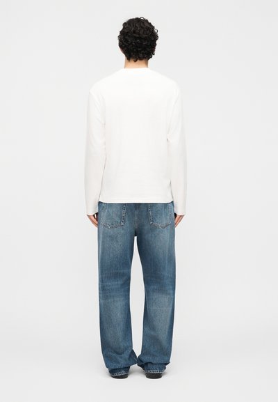 Άνδρας με κατσαρά μαλλιά φοράει λευκό πουκάμισο με μακριά μανίκια και φαρδιά μπλε τζην, στέκεται αντικριστός σε απλό λευκό φόντο.