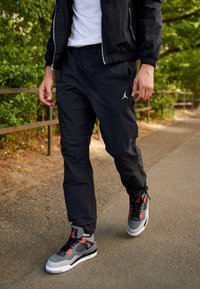 Černé joggingové kalhoty s elastickými manžetami, spárované se šedými a červenými teniskami s síťovinovými detaily a bílými podrážkami, umístěné na přírodním pozadí.