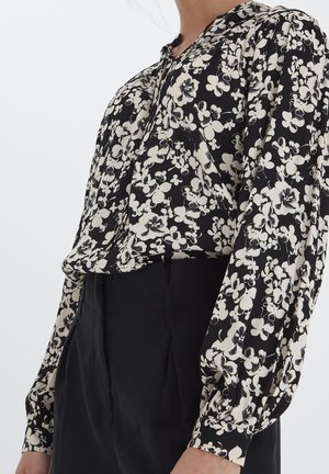Zwarte blouse met wit bloemenpatroon; lichte stof, lange pofmouwen, knoopsluiting aan de voorkant en een relaxed fit.