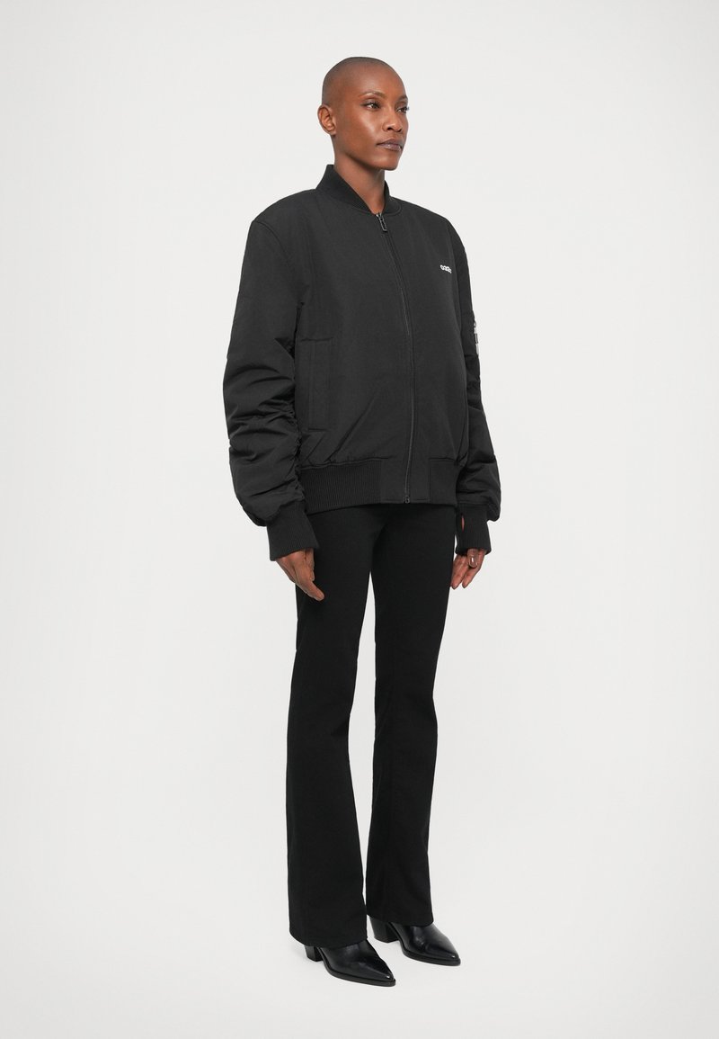 Person standing sideways wearing a black zip-up bomber jacket, black flared pants, and black pointed boots on a plain white background.