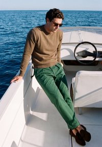 Homme portant des lunettes de soleil, un pull marron et un pantalon vert assis sur un bateau blanc près du volant avec l'océan en arrière-plan.