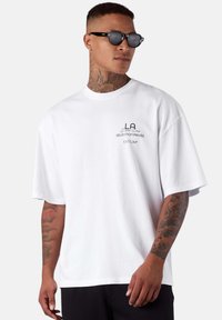 White cotton t-shirt with a loose fit; features a small black logo and text on the left chest. The model wears black sunglasses.
