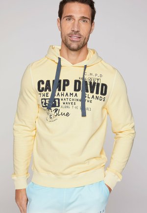 Sudadera amarilla con capucha ajustable, presenta texto y gráficos en negro en el frontal. Puños y dobladillo acanalados, con un corte holgado.