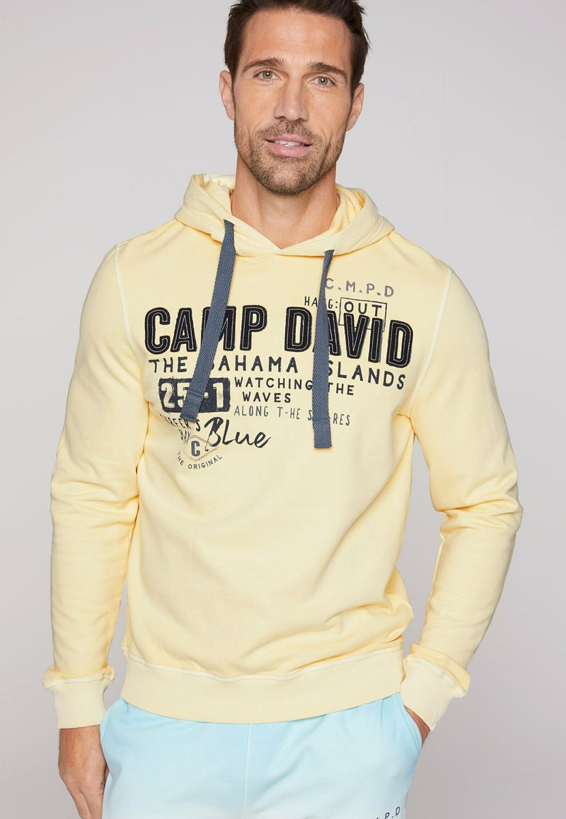 Sudadera amarilla con capucha ajustable, presenta texto y gráficos en negro en el frontal. Puños y dobladillo acanalados, con un corte holgado.