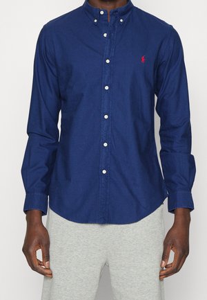 Mann trägt ein dunkelblaues Hemd mit Knöpfen und langen Ärmeln, einem roten Logo auf der Brust und hellgraue Hose.